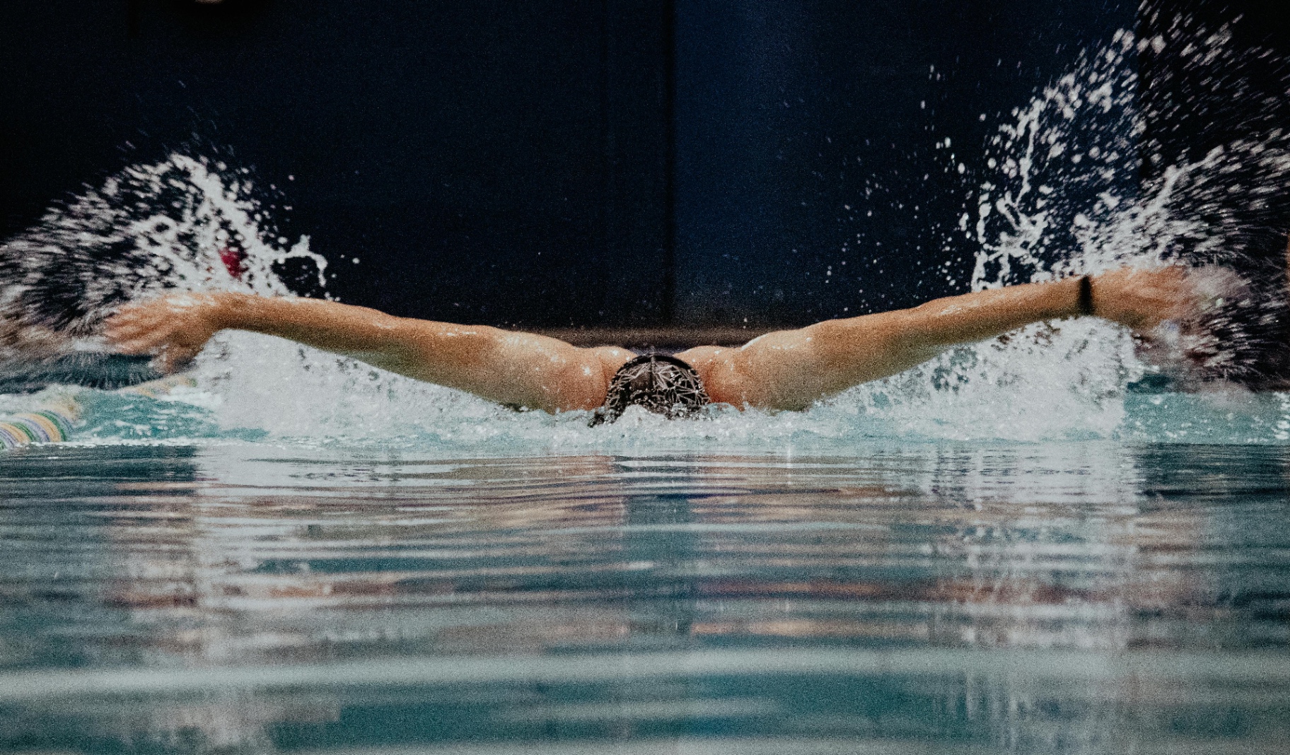 A triathlete swimming front crawl in a pool, arms extended in the recovery phase, showing the overhead reach that loads the rotator cuff