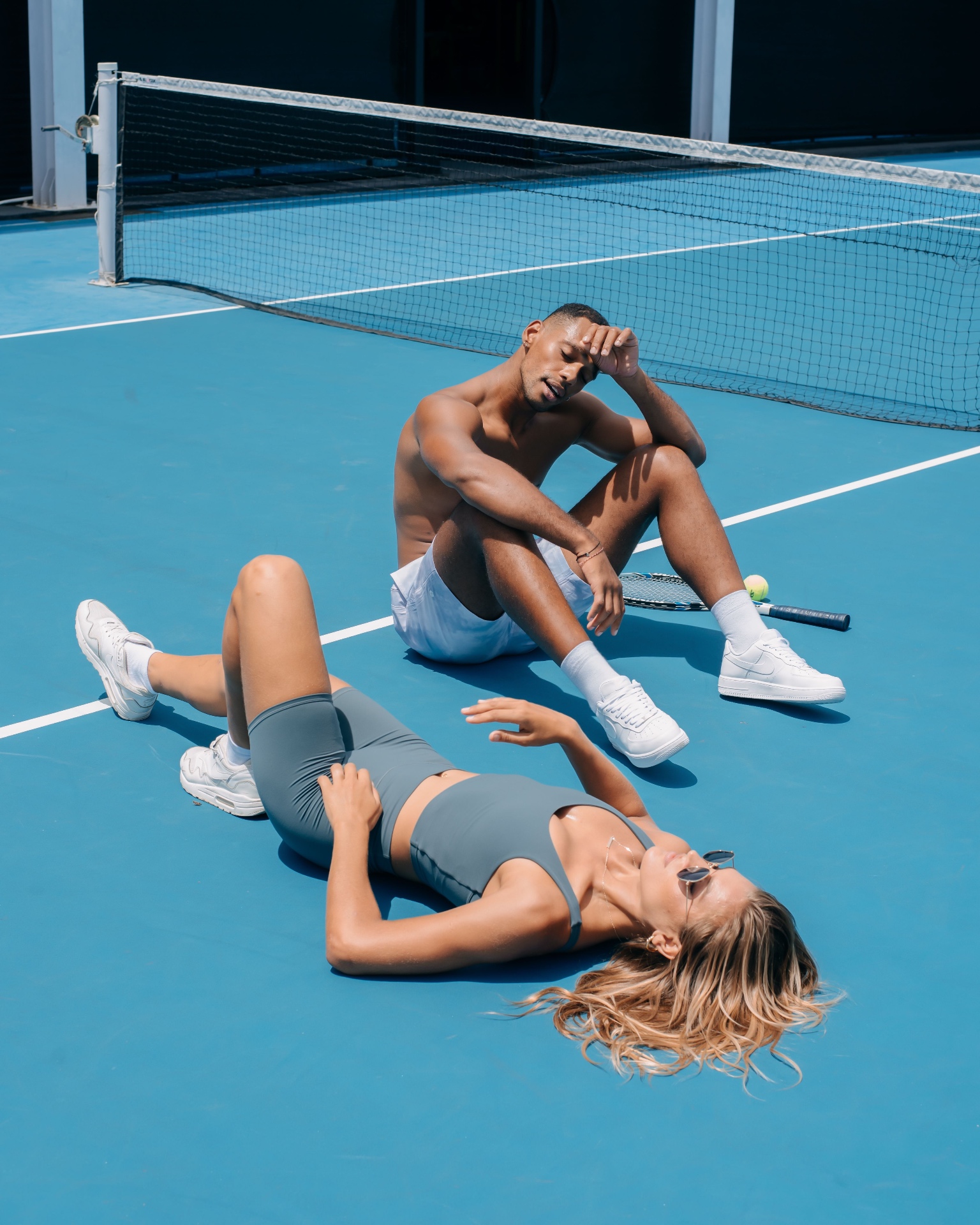 Two players resting on a blue padel/tennis court after an intense match, one seated by the net and one lying on the court — illustrating the physical demands of racket sports on the Costa del Sol